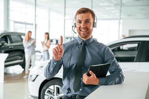 Manager, is in the headphones and with notepad. Handsome car dealership worker is with electric vehicle photo