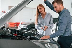 Showing engine under the hood. Man in formal clothes is with woman customer with the electric car photo