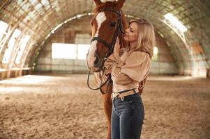 In, jeans and creamy ,colored clothes. Beautiful young woman is with horse indoors photo
