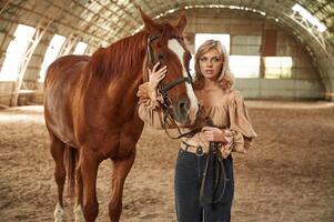 In jeans, and creamy colored clothes. Beautiful young woman is with horse indoors photo