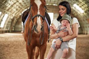 Holding ,kid, in hands. Gorgeous woman with her little baby son is with horse indoors photo
