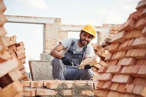 Sitting, and holding brick and little showel. Handsome Indian man is on the construction site photo