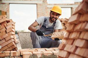 Sitting and, holding brick and little showel. Handsome Indian man is on the construction site photo