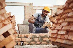 Sitting and holding brick and little showel. Handsome Indian man is on the construction site photo
