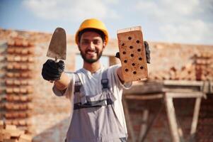 Showing and holding, little ,shovel, and brick. Handsome Indian man is on the construction site photo