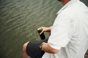 Close up view ,of man that is sitting by the water with beer in hands photo