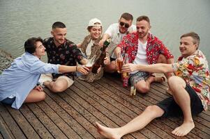 Celebrating,, having fun.. Group, of friends are sitting near the lake with beer in hands photo