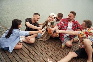 Celebrating, having fun. Group ,of ,friends, are sitting near the lake with beer in hands photo