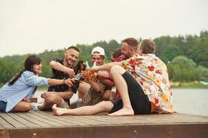 On the wooden floor. Group, of ,friends are sitting near the lake with beer in hands photo