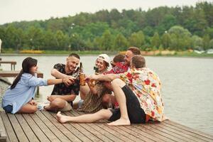 On the wooden, floor. Group of friends are sitting near the lake with beer in hands photo