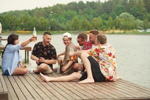 On the, wooden floor. Group of friends are sitting near the lake with beer in hands photo