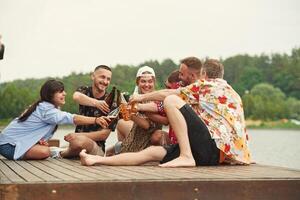 On, the wooden floor. Group of friends are sitting near the lake with beer in hands photo