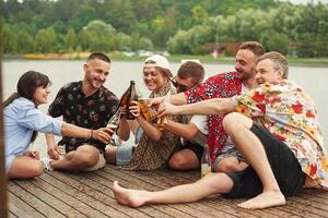 On the, wooden, floor. Group of friends are sitting near the lake with beer in hands photo