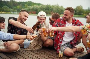 Celebrating, having, fun. Group of friends are sitting near the lake with beer in hands photo