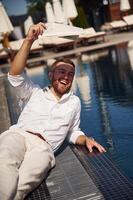 Covering himself by newspaper from bright sunlight. Handsome man in formal clothes is in the pool photo