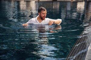 Checking ,the time, in the water. Handsome man in formal clothes is in the pool photo