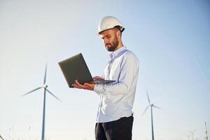 Standing and using laptop.. Service, engineer is on the field with windmills photo