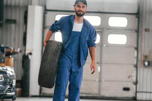 Front view, standing, and ,holding tire. Auto mechanic working in garage. Repair service photo
