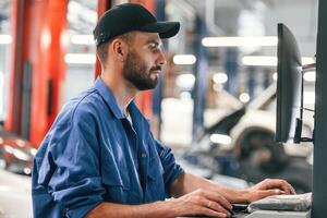 In black ,cap. Using, computer. Auto mechanic working in garage. Repair service photo