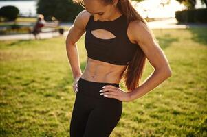 Showing abs. Beautiful young woman in sportive clothes is in the park at sunny daytime photo