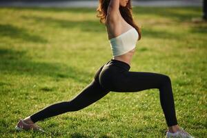 On green grass, stretching the legs. Beautiful young woman in sportive clothes is in the park at sunny daytime photo