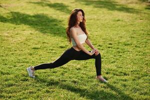 On green grass, stretching ,the, legs. Beautiful young woman in sportive clothes is in the park at sunny daytime photo