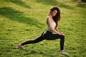 On green grass, stretching the, legs. Beautiful young woman in sportive clothes is in the park at sunny daytime photo