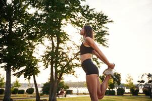 Exercises, against tree and sky... Beautiful young woman in sportive clothes is in the park at sunny daytime photo