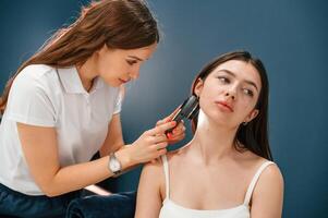 Against blue wall. Beauty salon worker is examining skin of the woman by using a device with light and lens photo