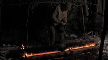 A man sits by a glowing campfire under a makeshift shelter in the forest at night, illuminated by a headlamp. The scene captures the essence of wilderness survival. video