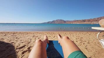 A relaxing beach scene captured from a first-person perspective. The image shows legs stretched out on a lounge chair or mat, with golden sand in the foreground. video