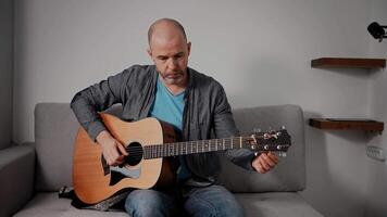 A middle-aged man sits on a gray couch, tuning an acoustic guitar with focused attention. The room has a minimalist decor, keeping the focus on the man and his instrument in a relaxed setting. video