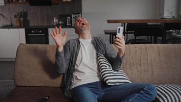 A very cheerful middle-aged man laughs heartily while chatting on a call using his smartphone. He is sitting at home on the sofa in the living room. An emotional man with a great sense of humor video