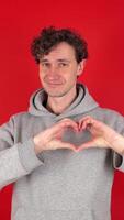 young adult, cheerful, curly haired brunette man in gray hoodie shows shape of heart frame with two hands in front. Isolated on red background of studio shot. Concept - love sympathy, romance, voting video