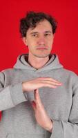 young adult curly serious man holds hands in front of shows sign timeout gesture. wearing grey hoodie. isolated red background studio shot. Concept - pause, break, rest, vacation video