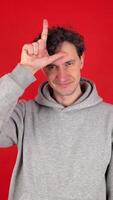 young adult curly serious man raises hand making loser gesture, L sign on forehead, teasing, accusing for defeat, expressing disrespect. Isolated red background studio shot. Concept - you lose. video