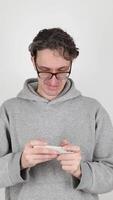 young man, geek in gray hoodie, glasses actively playing and winning computer game on mobile phone by giving thumbs up at the end. isolated on white background. Concept - mobile applications, winner video