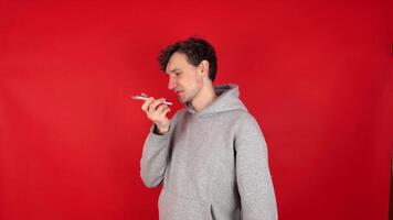 curly man in gray hoodie recording voice message on smartphone, talking speakerphone. Isolated red background studio shot. concept - communication, technology, social media. video