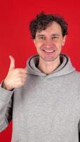 Young adult cheerful curly man showing in front thumb up hand sign like gesture, looking smiling at camera. Isolated red background studio shot. Concept - happy, voting, approval, social media video