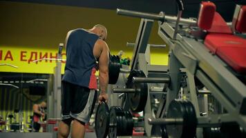 bald athletic man beard focuses on leg exercises in gym setting during evening. He adjusts weights on machine showcasing his dedication fitness video