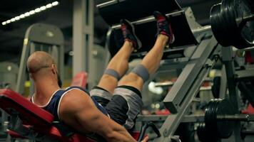 bald athletic man beard is engaged in leg exercises using weight machine in well equipped gym. He appears focused and determined during his midday workout video