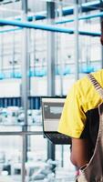 Vertical Employee using laptop in smart factory, collecting and analyzing data. African american woman using tracking software in industry 4.0 industrial plant equipped with advanced sensors, camera A video