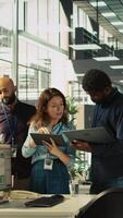 Vertical Multiethnic coworkers looking over revenue figures on tablet in diverse office. Multiracial colleagues doing brainstorming, comparing company statistical numbers, working hard to finish job, camera B video