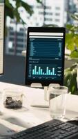 Vertical Modern workspace with analytical data dashboards displayed on devices in bright office environment. PC screen on desk with financial statistics graphs next to potted plants, jib down shot video