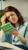 Vertical Stressed woman in office feeling painful headache while working, suffering burnout, reading medication box instructions. Employee having migraine after working overtime to finish project, camera B video