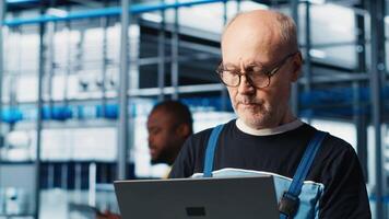 Smart factory senior engineer uses laptop to oversee operation of robotic arms and conveyor systems. Experienced industry 4.0 plant technician monitors system performance using notebook, camera A video