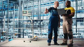 Senior engineer talking with intern in smart factory, collecting and analyzing data. Experienced technician helping trainee use tracking software in industrial plant equipped with sensors, camera A video