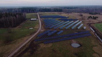 Rows of solar panels in an open field with a dirt road, small building, pond, and bare trees under a cloudy sky, surrounded by grassland and forest. video