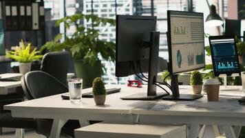 Business graphs and performance metrics on monitors in workplace, handheld camera shot. Office desk setup in bright corporate environment with monitors displaying data analytics, panning shot video