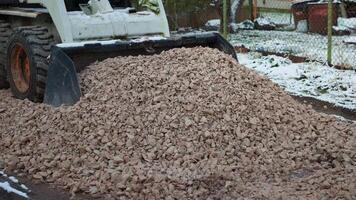 A small construction vehicle distributes crushed stones on a pathway in a winter setting. Snow patches, trees, and a chain link fence are visible. video
